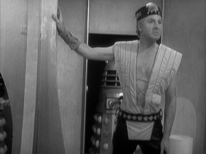 Temmossus, leader of the Thals, stands in the Dalek city. He is an older blond man wearing a puffy vest, no shirt, and groovy trousers. Two Daleks lurk behind him, unseen.